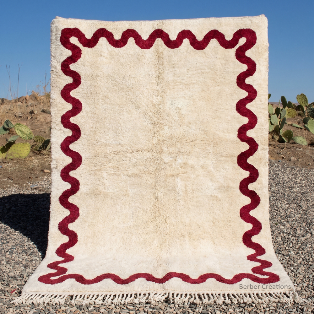 The Crimson Ripple authentic burgundy wavy-border Moroccan wool rug set against a natural landscape, featuring a large, plush cream field and traditional hand-knotted wool fringes.