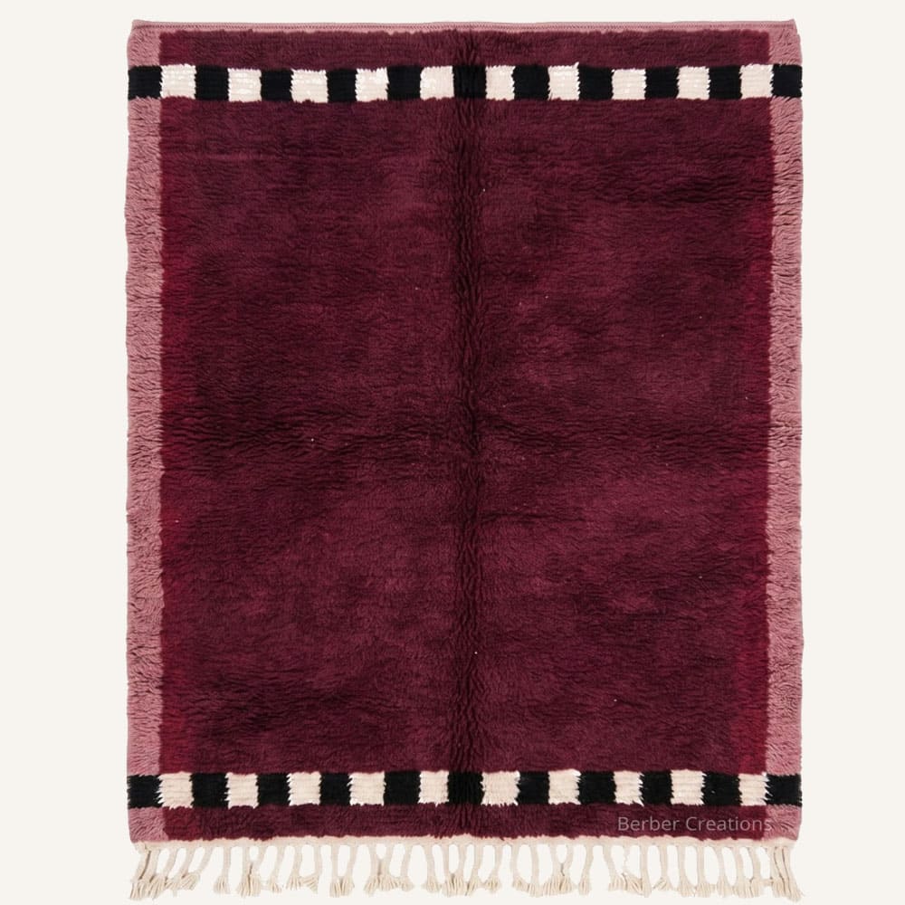 Tazoughart handmade Moroccan Berber wool rug featuring a rich deep burgundy center framed by dusty rose side selvedges and black and cream checkered end bands with braided fringe.