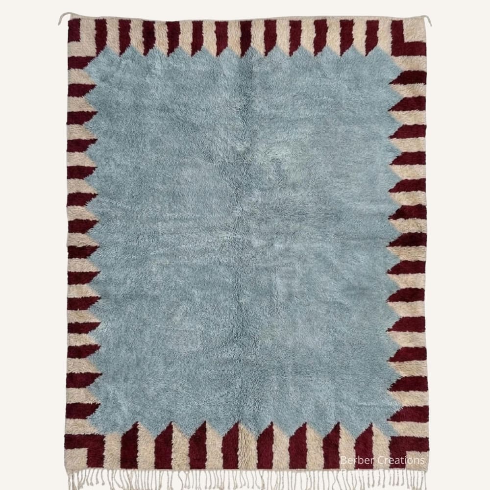 Tisi handmade Moroccan Berber wool rug featuring a central plush dusty slate blue field framed by jagged burgundy sawtooth points against a warm oatmeal beige outer border